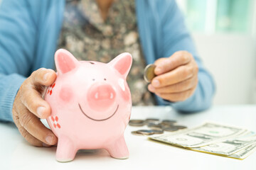 Asian elderly woman putting coin into pink piggy bank for saving money and insurance, poverty,...