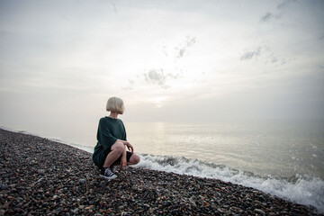 Sitting young girl with short blonde bob hair and green dress on cold sea beach