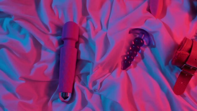 set of woman sex toys for masturbation and orgasm on sheet on bed. Female vibrator, vacuum stimulator, butt plug and BDSM handcuffs in neon lighting