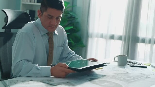 Professional engineer looking at phone while working on tablet with green screen at meeting table with turbine engine or jet engine sketch design and various metal gear scatter around. Alimentation.