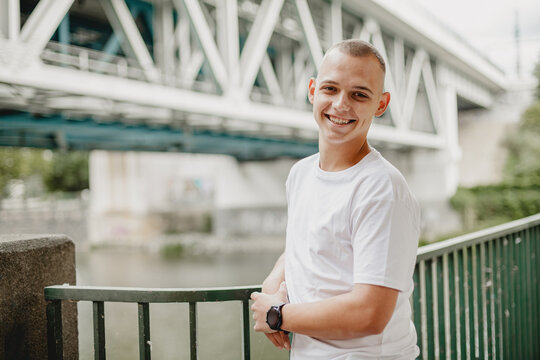 A man is smiling and posing for a picture in front of a bridge. The bridge is tall and spans across a body of water. The man is wearing a white shirt and a watch