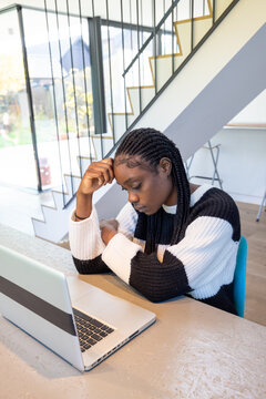 An image portraying a young woman in deep concentration while working on her laptop. Her braided hair and contemporary outfit, along with the stylish home setting with staircases in the background