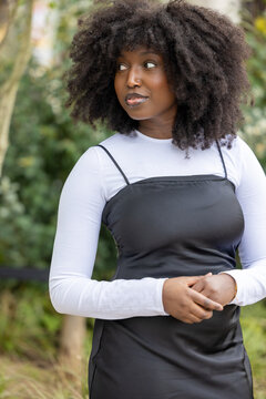 A natural-light portrait of a contemplative woman dressed in a modern layered ensemble, with a black slip dress over a white long-sleeve shirt. Her expressive afro hairstyle and the soft outdoor