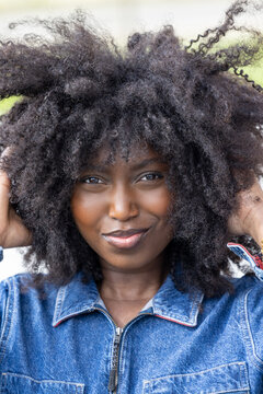 A close-up portrait of a vivacious woman with a playful afro hairstyle, giving a subtle, confident smile to the camera. She sports a denim jacket that adds a casual yet edgy touch to her look