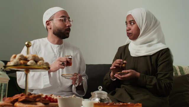 Happy young Muslim woman in hijab looking at her husband in white kandora and skullcap while both sitting on couch, having hot tea and chatting by breakfast