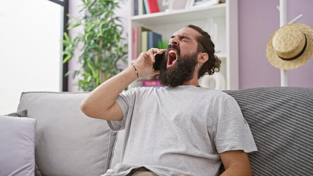 A young hispanic man with a beard bostezando while on a cellphone at his stylish living room, wearing a casual t-shirt and touching his beard.