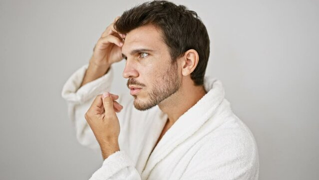 Pensive hispanic man in white bathrobe against isolated background poses with hand on head, looking away thoughtfully.