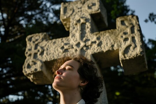 person standing with eyes closed before a large stone cross