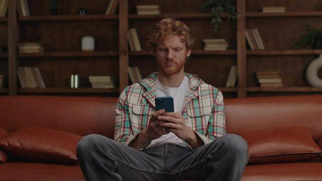 Relaxed young man with ginger hair browsing his phone while sitting on a leather sofa in a stylish interior.