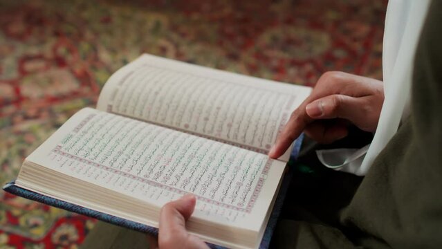 Hands of unrecognizable Muslim woman in white hijab holding open Holy Quran and reading verses of surah while sitting in front of camera and turning pages of book