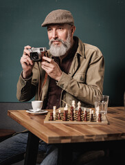Portrait d'un homme photographe quinquagénaiore agé senior avec une barbe et un béret tenant un appareil photo argentique et assis à une table avec un jeux d'échec