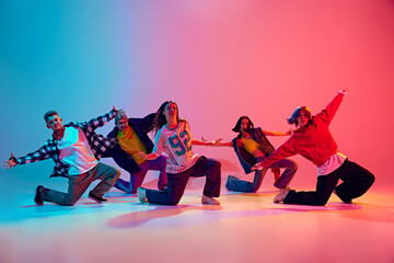 Dynamic squad of young dancers performing their routine in neon light against gradient colorful studio background. Concept of hobby, sport, fashion and style, action, youth culture, music and dance.