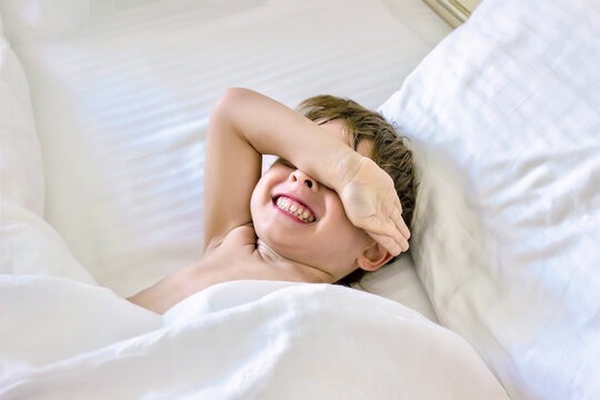 Adorable little toddler boy trying to sleep in white bed. Child is going to have a rest or a nap. Kid is smiling, hiding himself by his hand. Soft focus.