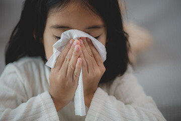 An unhealthy kid blowing their nose into a tissue, a Child suffering from running nose or sneezing, A girl catching a cold when the season change, a childhood wiping nose with a tissue