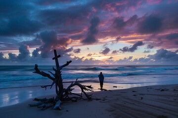 person at sunrise on a beach, praying near driftwood cross - obrazy, fototapety, plakaty