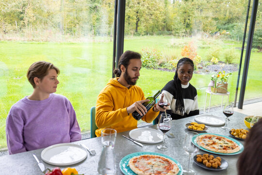 A cheerful gathering of friends at a dining table, enjoying an array of appetizing foods and wine in a light-filled greenhouse setting. The lush green backdrop seen through the glass walls adds a