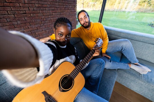In this inviting scene, two friends share a moment of music and companionship. A woman, extending her arm for a selfie, holds a guitar, while a man beside her relaxes on the couch. The sunlit room and