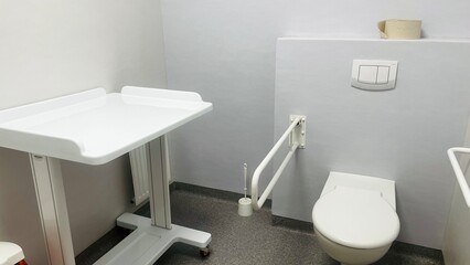 white toilet with metal grab bars and changing table in a toilet in a hospital with gray walls