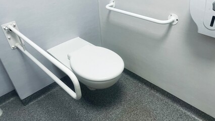 white toilet with metal grab bars in a toilet in a hospital with gray walls. side view