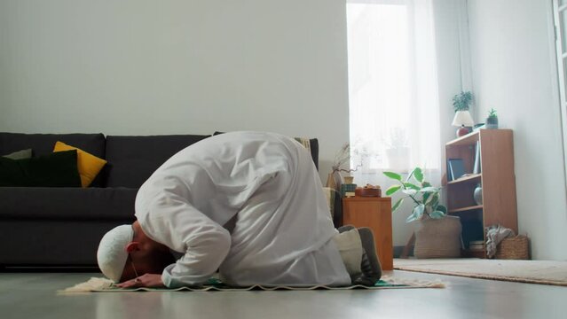 Young Muslim man in white skullcap and kandora standing on his knees on the floor of living room, bending forwards and touching rug by head during prayer
