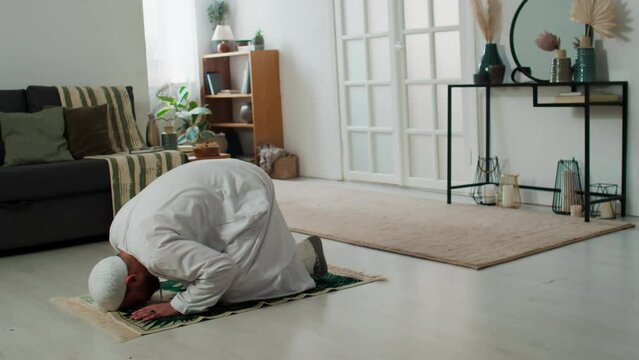 Knelt young man in traditional Islamic skullcap and thobe bending forwards and touching rug by head during prayer on the floor of living room
