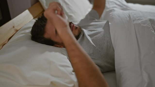 A relaxed young hispanic man with a beard lying on a bed indoors, depicting a comfortable home bedroom setting.