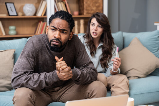 Unhappy young woman sitting on sofa with pregnancy test, African American husband screaming at her, indoors. Millennial family having conflict over unexpected childbearing, argue over results