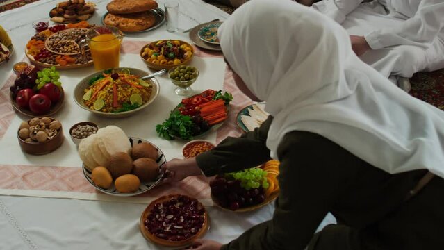 High angle of young Muslim woman in white hijab standing on knees in front of tablecloth and putting plate with fresh grapes next to sweet pie, buns and other national food prepared for dinner