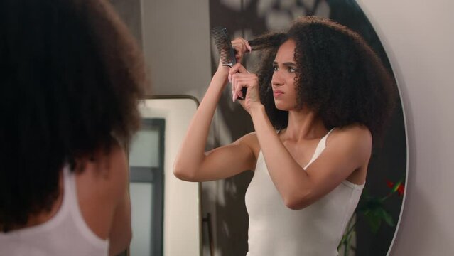 Morning beauty routine procedure healthy African American girl woman female at home bathroom looking at mirror haircare problem upset suffering dry mess curly hair trying to brush tangled hairdo