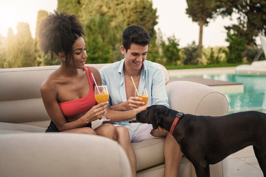 Multiethnic Couple with refreshing drinks by pool, enjoying company of pet dog - tranquil leisure, summer vibes, poolside relaxation. - Powered by Adobe