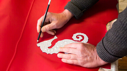 A tailor traces a stencil of a seahorse on a red canvas with his hand and a pencil
