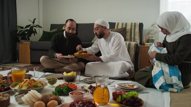 Young Muslim man in white thobe and skullcap putting homemade food on plate held by his twin brother during festive dinner while woman in hijab lulling baby