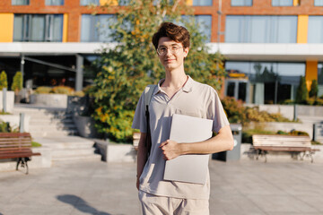 A young man stands in front of a building with a laptop in hand