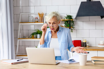 beautiful elderly senior woman in cosy kitchen works on laptop and speaking by phone, beautiful gray haired woman works online, pays bills, studies and communicates, concept of active old age