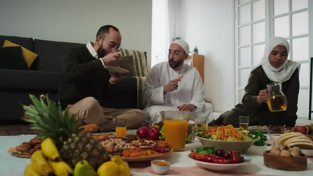 Young woman in hijab pouring juice or lemonade in glass and drinking it while her husband and his twin brother eating homemade baked potatoes and chatting by dinner