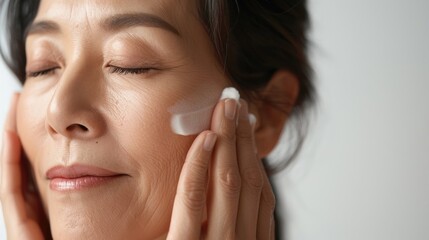 A woman with her eyes closed applying a white cream to her face with both hands focusing on her skin care routine.