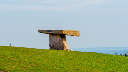 A solitary ancient rock table stands against a backdrop of clear blue sky atop a lush green hillside, with a calm sea visible in the distance.