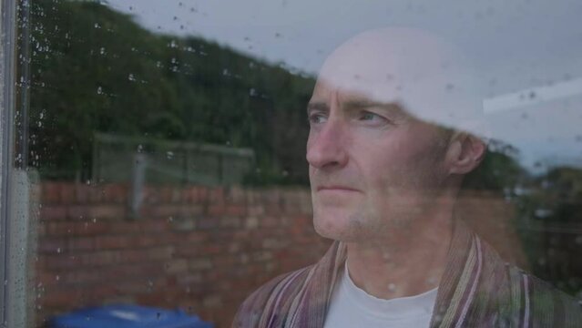 Male looks out of house window depressed and bored. Middle aged man wearing a dressing gown. Male mental health, self isolation. Cost of living crisis. Midlife crisis. 