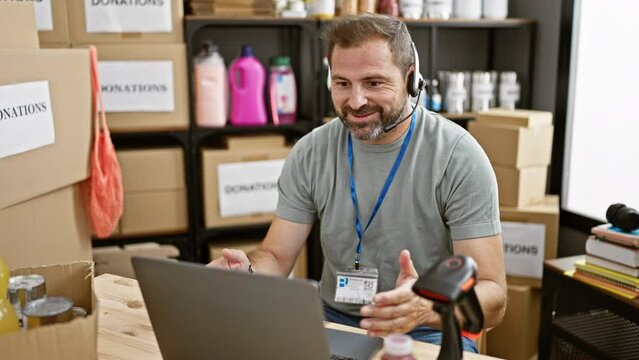 Middle-aged man with beard working on laptop in donation center warehouse, expressing various emotions.