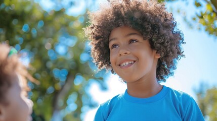 A young child with curly hair wearing a blue t-shirt smiling and looking up towards the sky with a blurred background of trees and a clear blue sky.