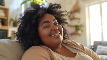 A joyful woman with curly hair wearing a beige top smiling broadly and sitting comfortably on a couch in a brightly lit room with plants and a window in the background. - obrazy, fototapety, plakaty