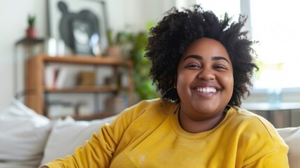 Fototapeta premium A joyful woman with curly hair wearing a yellow top sitting on a couch in a cozy room with plants and a painting in the background.