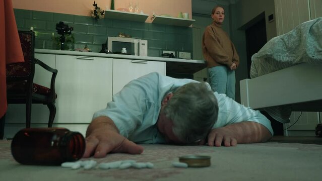Elderly man lies on floor, probably having heart attack and trying to reach for pills, young woman - daughter or carer looks on him cold blood.