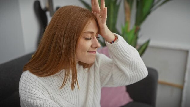 Oops! young redhead woman, surprised at home wearing sweater, hand on head remembering forgotten mistake - a foolish blunder hitting her memory