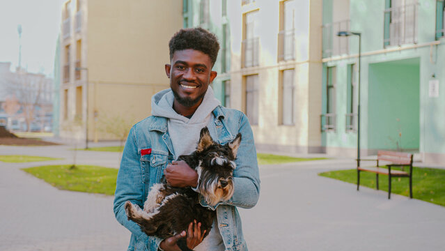 African american young man with smile on face embracing purebred pug dog while standing at urban area. Cute scottish terrier relaxing on owner s hands after long walk on city street.