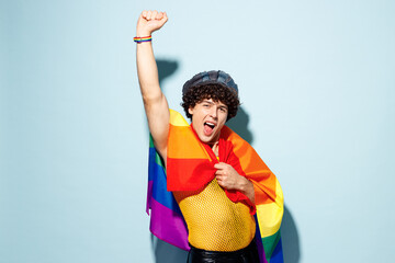 Young happy gay Latin man wears mesh tank top hat clothes wrapped in striped rainbow flag raise up...