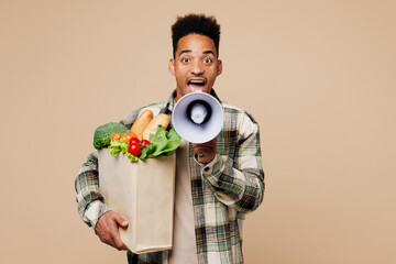 Young man wear grey shirt hold paper bag takeaway mock up with food products scream in megaphone announces discounts sale isolated on plain beige background. Delivery service from shop or restaurant.