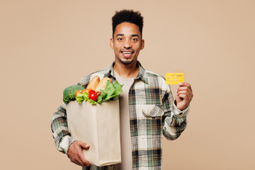 Young smiling man wears grey shirt hold paper bag for takeaway mock up with food products, credit bank card isolated on plain pastel light beige background. Delivery service from shop or restaurant.
