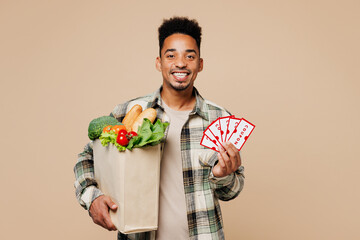 Young fun man wear grey shirt hold paper bag for takeaway mock up with food products, show many coupon cards isolated on plain pastel light beige background. Delivery service from shop or restaurant.