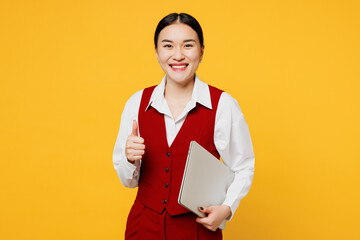 Young corporate lawyer employee business IT woman of Asian ethnicity wear red vest shirt work at office hold closed laptop pc computer show thumb up isolated on plain yellow background Career concept